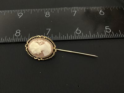 Sale Photo Thumbnail #172: A vintage gold-tone stickpin featuring an oval carved shell cameo depicting a woman in profile. The cameo is set in a decorative ornate filigree and rope-edge frame. The back of the setting features detailed scrollwork and filigree design. Total length me