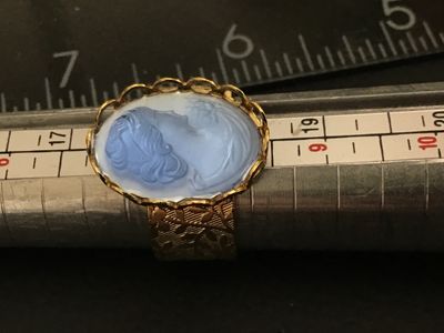 Sale Photo Thumbnail #194: A vintage costume jewelry ring featuring a blue glass molded cameo depicting a classic portrait profile. Set in a scalloped bezel with a wide gold-tone band decorated with an etched floral leaf pattern. Ring measures approximately size 8.5 to 8.75 on a ri
