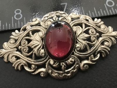Sale Photo Thumbnail #70: An ornate vintage silver-tone metal brooch featuring detailed filigree and scrollwork, set with a central oval dark purple or garnet-colored glass cabochon stone. The brooch measures approximately 2.5 inches in length. The back features a standard pin cla