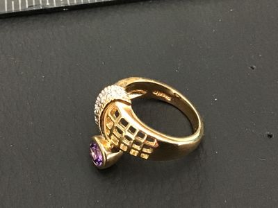 Sale Photo Thumbnail #242: A distinctive gold-tone statement ring featuring a bypass-style lattice/grid design, accent stones along the band, and a bezel-set purple round stone. Shown on a ring mandrel measuring approximately size 6 to 6.5. Good condition with light surface wear.