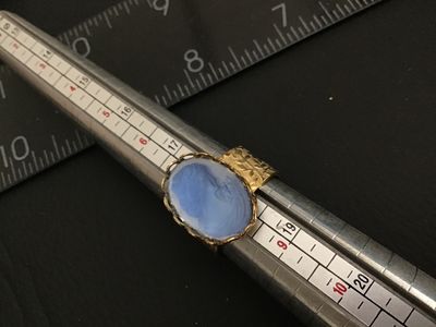 Sale Photo Thumbnail #196: A vintage costume jewelry ring featuring a blue glass molded cameo depicting a classic portrait profile. Set in a scalloped bezel with a wide gold-tone band decorated with an etched floral leaf pattern. Ring measures approximately size 8.5 to 8.75 on a ri