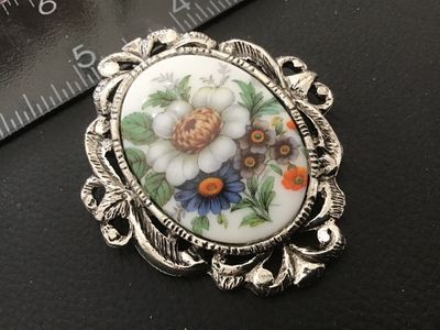Sale Photo Thumbnail #106: Vintage oval brooch featuring a hand-painted or printed porcelain/ceramic insert with a colorful floral motif including white, blue, and orange flowers. Set in an ornate, openwork silver-tone metal frame with textured scroll and leaf detailing. Includes b