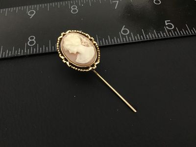 Sale Photo Thumbnail #169: A vintage gold-tone stickpin featuring an oval carved shell cameo depicting a woman in profile. The cameo is set in a decorative ornate filigree and rope-edge frame. The back of the setting features detailed scrollwork and filigree design. Total length me