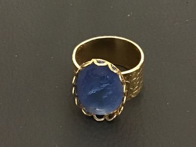 Sale Photo Thumbnail #200: A vintage costume jewelry ring featuring a blue glass molded cameo depicting a classic portrait profile. Set in a scalloped bezel with a wide gold-tone band decorated with an etched floral leaf pattern. Ring measures approximately size 8.5 to 8.75 on a ri