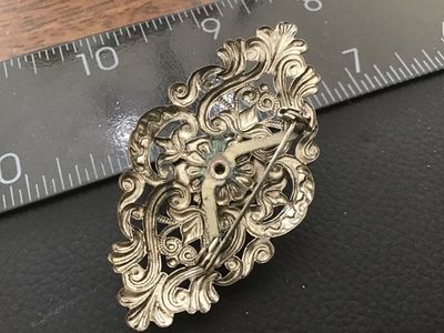 Sale Photo Thumbnail #71: An ornate vintage silver-tone metal brooch featuring detailed filigree and scrollwork, set with a central oval dark purple or garnet-colored glass cabochon stone. The brooch measures approximately 2.5 inches in length. The back features a standard pin cla