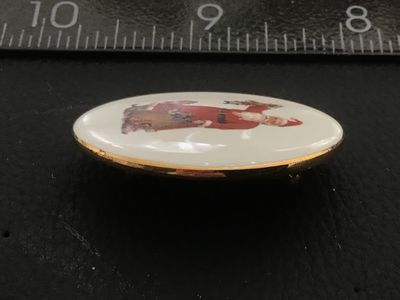 Sale Photo Thumbnail #52: Oval porcelain cabochon brooch featuring a classic vintage image of Santa Claus carrying a sack of toys. Framed in gold-tone metal with a pinback closure. Measures approximately 1.5 inches wide. Shows minor signs of age and wear on the back rim, pin mecha