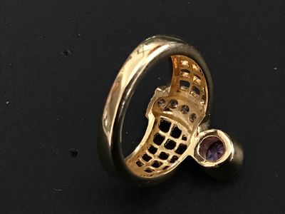 Sale Photo Thumbnail #244: A distinctive gold-tone statement ring featuring a bypass-style lattice/grid design, accent stones along the band, and a bezel-set purple round stone. Shown on a ring mandrel measuring approximately size 6 to 6.5. Good condition with light surface wear.
