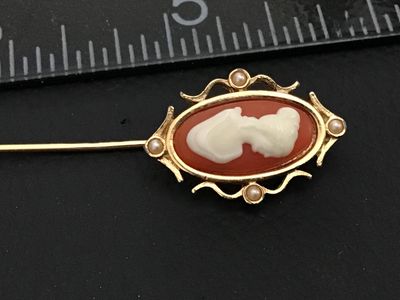Sale Photo Thumbnail #173: Vintage gold-tone stick pin featuring an oval faux-cameo with a white profile against a terracotta-red background, surrounded by a scrolled frame accent with four small faux pearls. Signed "© SARAH COV" on the back. Measures approximately 3 inches in tot