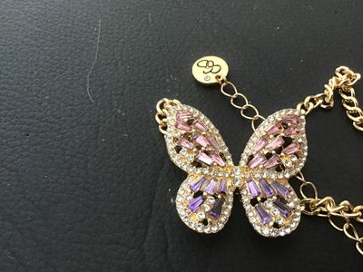 Sale Photo Thumbnail #105: Gold-tone chain necklace featuring a stylized butterfly pendant set with purple, pink, and clear rhinestones. Features a logo hang tag at the clasp extender. Measures approximately 11 inches in total laid-flat length including the pendant. In good pre-own