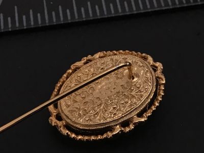 Sale Photo Thumbnail #170: A vintage gold-tone stickpin featuring an oval carved shell cameo depicting a woman in profile. The cameo is set in a decorative ornate filigree and rope-edge frame. The back of the setting features detailed scrollwork and filigree design. Total length me