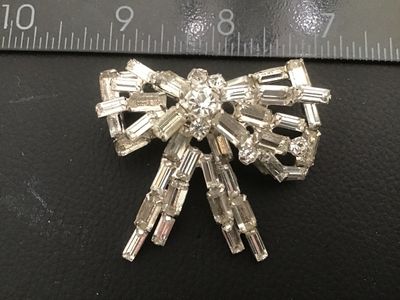 Sale Photo Thumbnail #53: A vintage silver-tone costume jewelry brooch in a dimensional bow design, set with baguette and round cut clear rhinestones. Features a pin stem and roll-over safety clasp on the reverse. Measures approximately 2 inches wide by 2 inches long. Good vintage