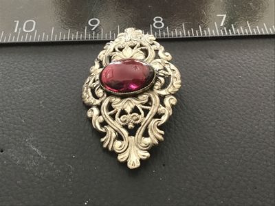 Sale Photo Thumbnail #72: An ornate vintage silver-tone metal brooch featuring detailed filigree and scrollwork, set with a central oval dark purple or garnet-colored glass cabochon stone. The brooch measures approximately 2.5 inches in length. The back features a standard pin cla
