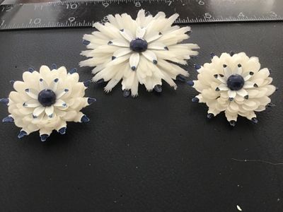 Sale Photo Thumbnail #134: Matching 3-piece vintage jewelry set featuring a large floral brooch pin and two clip-on earrings. Crafted from layered white celluloid petals with painted blue tips and a dark blue center cabochon accent. Brooch measures approximately 3.5 inches across. 