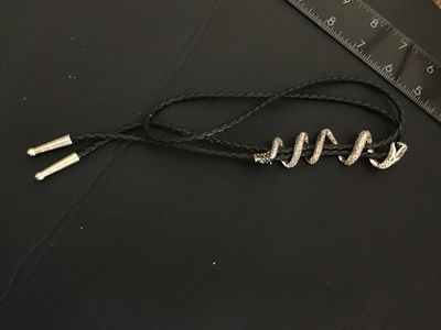 Sale Photo Thumbnail #256: Unique western bolo tie featuring a silver-tone coiling snake slide piece wrapped around a braided black leather/synthetic cord with silver-tone tips. Shows detailed scale texturing and open mouth snake design. Good condition with light wear.