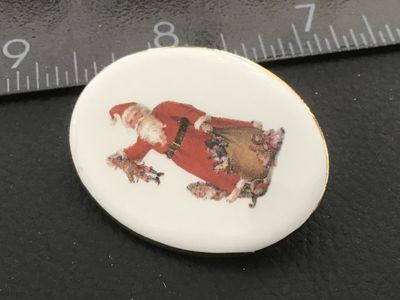 Sale Photo Thumbnail #50: Oval porcelain cabochon brooch featuring a classic vintage image of Santa Claus carrying a sack of toys. Framed in gold-tone metal with a pinback closure. Measures approximately 1.5 inches wide. Shows minor signs of age and wear on the back rim, pin mecha