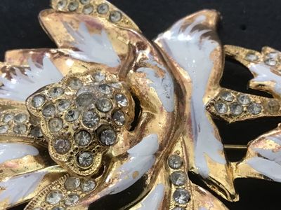 Sale Photo Thumbnail #114: Vintage gold-tone floral motif brooch pin featuring white enamel details and clear rhinestone accents. Shows noticeable surface wear, plating loss, enamel chipping, and age-related patina on the finish. The pin mechanism on the reverse is intact and funct
