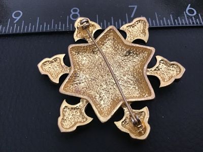 Sale Photo Thumbnail #147: Vintage ornate star / Maltese cross brooch crafted in a textured antique gold-tone metal. Detailed with raised beaded accents, geometric star patterning, and set with red and green rhinestones. Features a standard roll-over safety clasp pin back. Measures