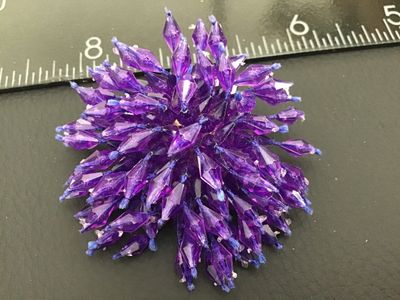 Sale Photo Thumbnail #266: A vintage starburst or pom-pom style brooch featuring faceted purple plastic beads wired/strung onto a gold-tone metal base with a rollover safety pin clasp. Measures approximately 2 inches across. Good pre-owned condition with light vintage wear to the m