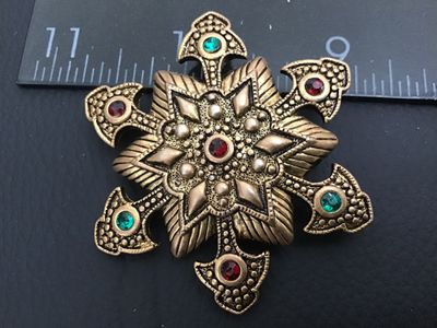 Sale Photo Thumbnail #148: Vintage ornate star / Maltese cross brooch crafted in a textured antique gold-tone metal. Detailed with raised beaded accents, geometric star patterning, and set with red and green rhinestones. Features a standard roll-over safety clasp pin back. Measures