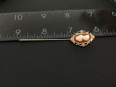 Sale Photo Thumbnail #177: Vintage gold-tone stick pin featuring an oval faux-cameo with a white profile against a terracotta-red background, surrounded by a scrolled frame accent with four small faux pearls. Signed "© SARAH COV" on the back. Measures approximately 3 inches in tot