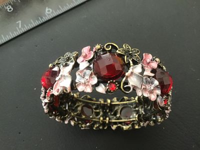 Sale Photo Thumbnail #87: A ornate stretch cuff bracelet featuring antiqued brass-tone metal set with large faceted red glass or acrylic stones, accent rhinestones, and delicate enameling on floral motifs in shades of pink, white, and grey. The bracelet features an elastic stretch