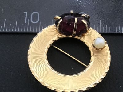 Sale Photo Thumbnail #57: Vintage circular gold-tone brooch featuring textured, engine-turned design details, accented with a single faux pearl and a prong-set faceted deep purple stone/glass element. Includes a standard pin mechanism on the reverse. Shows minor surface wear consi