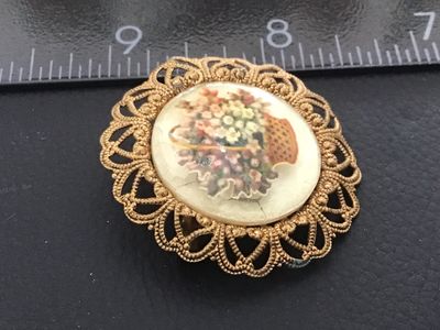 Sale Photo Thumbnail #94: Vintage round filigree brooch pin featuring a central cabochon decorated with a printed floral flower basket design. The gold-tone brass or alloy frame features ornate openwork filigree hearts and dots. Marked 'WESTERN GERMANY' on the back bar mechanism. 