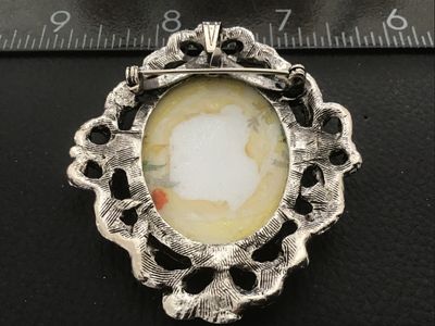 Sale Photo Thumbnail #108: Vintage oval brooch featuring a hand-painted or printed porcelain/ceramic insert with a colorful floral motif including white, blue, and orange flowers. Set in an ornate, openwork silver-tone metal frame with textured scroll and leaf detailing. Includes b