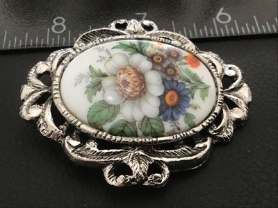 Sale Photo Thumbnail #107: Vintage oval brooch featuring a hand-painted or printed porcelain/ceramic insert with a colorful floral motif including white, blue, and orange flowers. Set in an ornate, openwork silver-tone metal frame with textured scroll and leaf detailing. Includes b