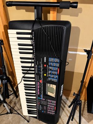 Sale Photo Thumbnail #7: Yamaha electronic keyboard stored vertically on a black stand, featuring built-in speakers, a digital display screen, and multi-colored control buttons.