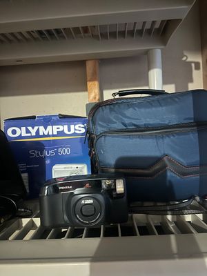 Sale Photo Thumbnail #32: Black Pentax IQZoom point-and-shoot 35mm film camera shown alongside a blue canvas camera bag and an Olympus Stylus 500 digital camera box.