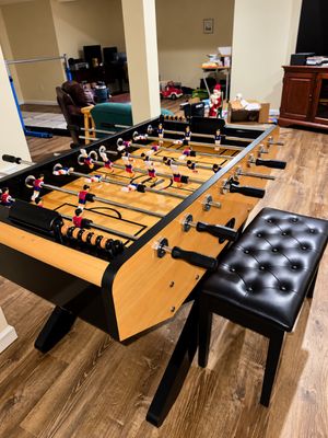 Sale Photo Thumbnail #10: Modern foosball table with a light wood grain finish, black trim, and telescoping rod handles, paired with a black tufted leatherette bench.