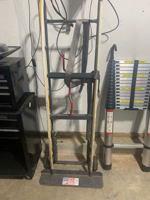 Sale Photo Thumbnail #13: Heavy-duty steel hand truck with an attached tie-down strap, shown next to a collapsible aluminum telescoping ladder.
