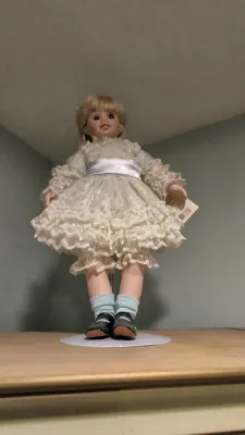 Vintage porcelain collector doll with blonde hair, wearing a ruffled lace dress with a satin sash and light blue socks.