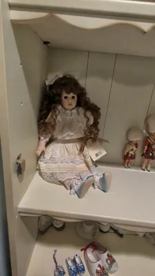 Vintage porcelain doll with curly brown hair in a lace dress, sitting on a shelf alongside smaller collectible figurines.