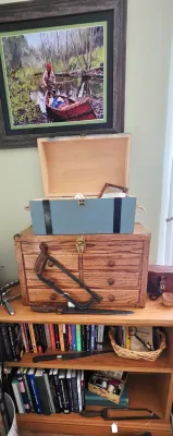 Sale Photo Thumbnail #5: Wooden bookcase filled with various collectibles, including a framed print of a canoeist, a wooden tool chest, a small blue box, vintage hand saws, a large knife, and assorted books.