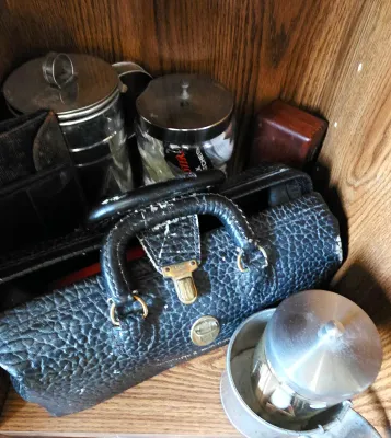 Sale Photo Thumbnail #18: Vintage textured black leather doctor's bag style pouch alongside glass canister jars and vintage metal items inside a wooden cabinet.