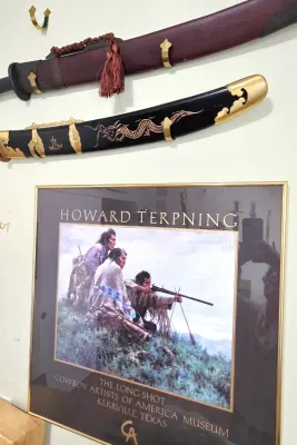 Sale Photo Thumbnail #57: Framed Howard Terpning art exhibition poster titled "The Long Shot" for the Cowboy Artists of America Museum, displayed below two decorative wall-mounted swords.