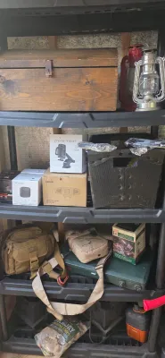 Sale Photo Thumbnail #25: Black plastic shelving unit stocked with vintage oil lanterns, a wooden chest, tactical bags, boxed electronics, and a green Coleman camping stove.