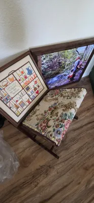 Sale Photo Thumbnail #50: Vintage tapestry upholstered footstool resting beside framed wall art, including a samper-style cross-stitch and a large landscape photograph.