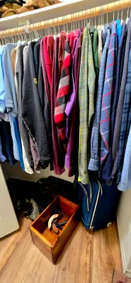 Sale Photo Thumbnail #53: Closet filled with men's clothing on hangers above a wooden crate and a navy blue rolling suitcase.
