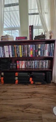Sale Photo Thumbnail #93: Dark wood three-shelf bookcase filled with hardback novels, a collection of DVDs, and stacked media cases along the bottom.