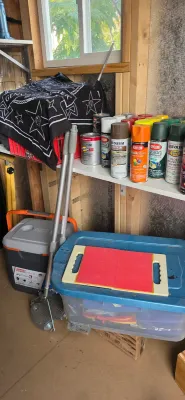 Sale Photo Thumbnail #34: Assortment of spray paint cans, paint tins, a Coleman cooler, blue storage tote, and lawn tools stored in a garage workspace.