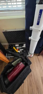 Sale Photo Thumbnail #98: Vixen A80Mf refractor telescope tube next to a red Orion telescope optical tube assembly, soft carrying cases, and an aluminum hard case filled with astronomical accessories.