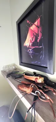 Sale Photo Thumbnail #13: Framed painting depicting a campsite at night, displayed above a shelf lined with various sheath knives, leather holsters, and glassware.