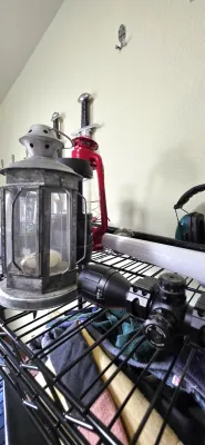 Sale Photo Thumbnail #67: Collection of outdoor and sporting goods including a vintage galvanized lantern, a red Dietz-style oil lamp, and a rifle scope resting on wire shelving.