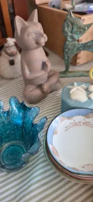 Sale Photo Thumbnail #86: Collection of cat-themed decorative items, including a meditating cat statue, a brass cat figurine, cat-face small ceramic dishes, and a blue ruffled art glass votive holder.