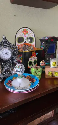 Sale Photo Thumbnail #105: Collection of Día de los Muertos and gothic decor items, including sugar skull plates, a painted light bulb nightlight, decorative tins, and an ornate table clock.
