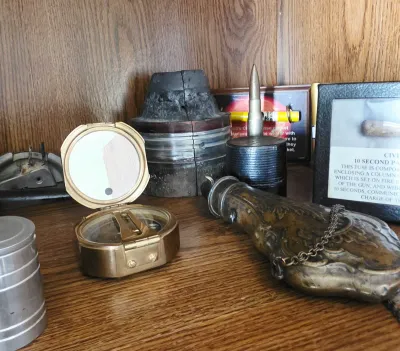 Sale Photo Thumbnail #15: Collection of vintage militaria and oddities, including an ornate metal powder flask, a brass pocket compass, inert ordnance, and framed historical displays.