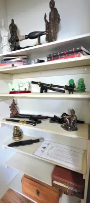 Sale Photo Thumbnail #68: Collection of Asian-themed decor items including brass and carved figurines, decorative swords, knives, books, and carved stone dogs on white shelving.